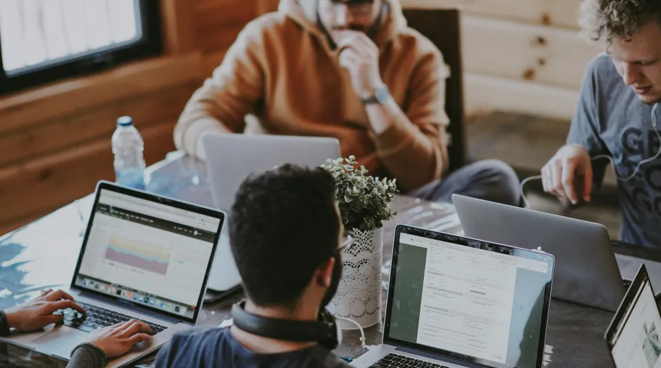 People collaborating indoors at a wooden table using laptops and technology in a cozy setting.