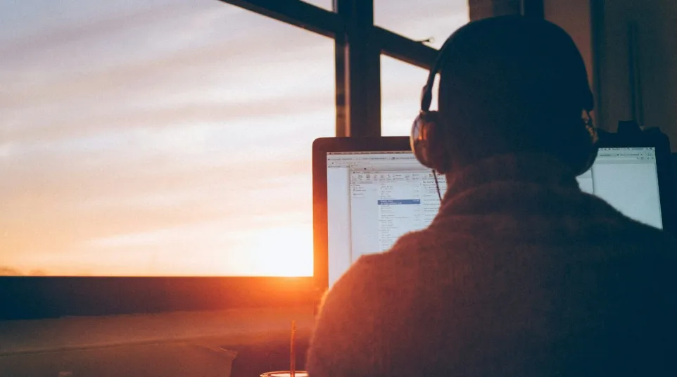 Person working on a computer at sunset, wearing headphones, with a view of the colorful sky.